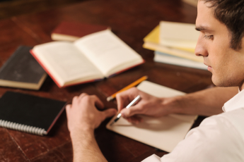 dark haired male student writing with fountain pen in notebook on desk with many open books