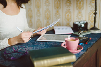woman reading some printed pages at a desk, with an ipad, cup of tea, notepad and some old books