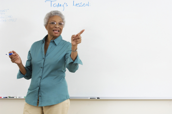 Female lecturer in blue shirt standing in front of a class with a blue whiteboard marker in one hand, and the phrase 'Today's Lesson' on the whiteboard behind her
